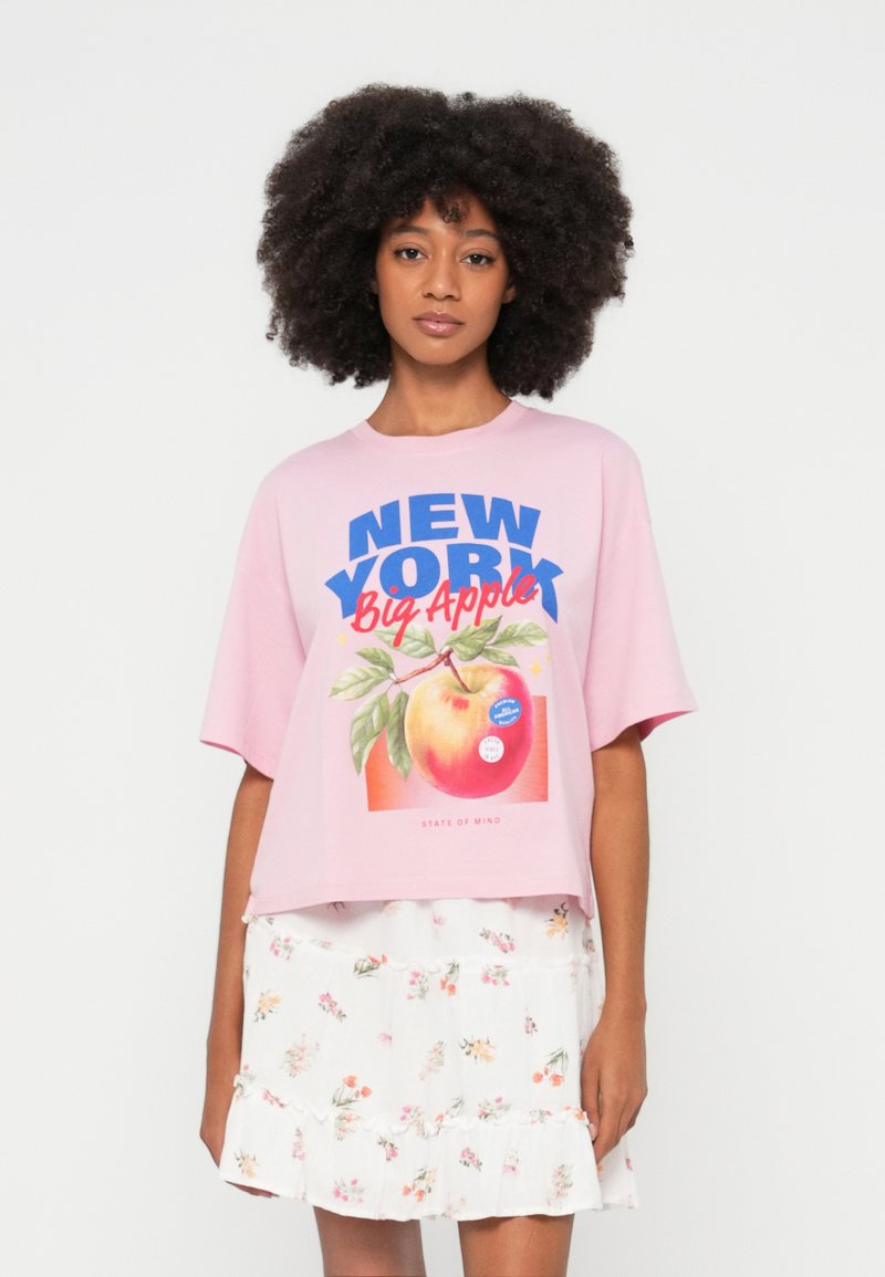Απαλό ροζ κοντό t-shirt με μπλε και κόκκινο γραφικό κείμενο που γράφει "New York Big Apple", με εικόνα ροδάκινου και λευκή φλοράλ φούστα.