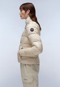 Beige pufferjack met hoge kraag, gequiltde textuur, zijzakken en een logo-opdruk op de linkerschouder. Soepele afwerking en slim fit ontwerp.