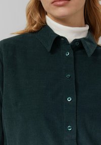 Chemise en velours côtelé vert foncé avec un col, présentant des boutons turquoise et un tissu texturé à côtes. Un col roulé blanc est visible en dessous.