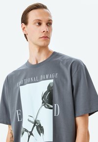 Camiseta de algodón gris con un gran gráfico de una rosa negra y el texto "DAÑO EMOCIONAL" y "ED" en blanco. Cuello redondo y corte holgado.