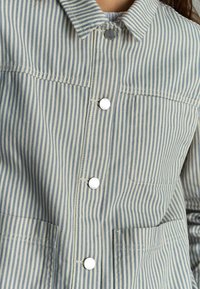 Gestreepte jas in lichtblauw en crème, met een kraag, knopen aan de voorkant en twee zakken aan de voorkant met witte knopen.