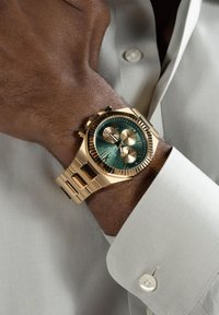 Gold-tone watch with a green textured dial, three sub-dials, and a metal band, positioned on a wrist with a light-colored, textured shirt.