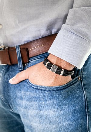 Black braided bracelet featuring silver accents, worn on a hand resting in a blue denim pocket, complemented by a brown leather belt.