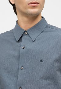 Κοντινό πλάνο ενός άνδρα που φορά μπλε πουκάμισο με κάθετες ρίγες, με γιακά και μικρό λογότυπο "cK" στο στήθος.