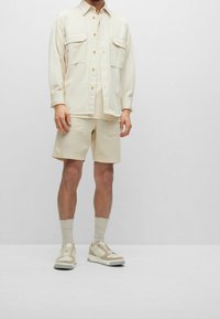 Muž v béžové košili s knoflíky a náprsními kapsami, odpovídajících šortkách, světlobéžových ponožkách a béžových teniskách, stojící proti jednoduchému pozadí.