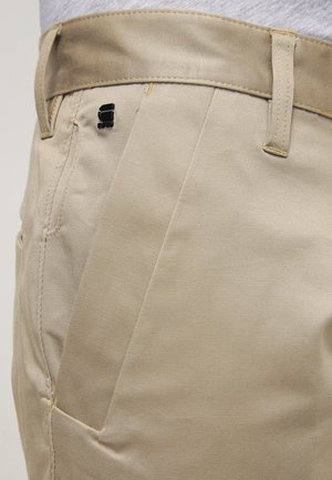 Beżowe spodnie z szlufkami na pasek, przednią kieszenią oraz małym czarnym logo przymocowanym przy szwie kieszeni w okolicy talii.