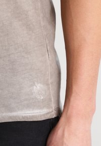 Šedé bavlněné tričko s kulatým výstřihem, krátkými rukávy a malým texturovaným detailem u spodního lemu. Černé kalhoty viditelné na noze modelky.