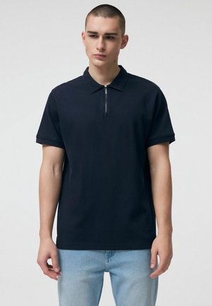 Młody mężczyzna o krótkich włosach, ubrany w czarną, krótka koszulkę polo z zamkiem i jasnoniebieskie dżinsy, stojący na jednolitym tle.