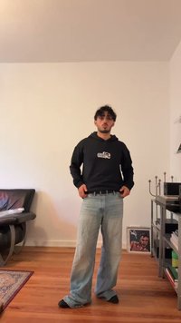 Giovane uomo con capelli ricci che indossa un felpa nera e jeans larghi di un blu chiaro, in piedi in un soggiorno minimalista con pavimento in legno.