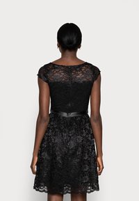 Robe en dentelle noire avec manches courtes type cap, corsage ajusté et jupe évasée. Dotée d'une ceinture en satin et de motifs floraux complexes.