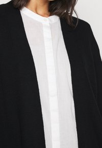 Svart cardigan med öppen frontdesign, parat med en vit, knapp-up, linnen skjorta. Cardiganen har en texturerad stickad finish.