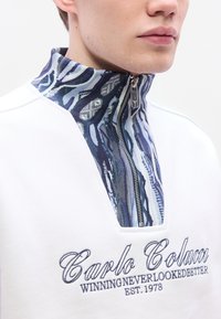 Λευκό πουλόβερ με ψηλό γιακά που διαθέτει μπλε σχέδιο στην επένδυση, κλείσιμο με μισή φερμουάρ και κεντημένο κείμενο "Carlo Colucci" στο μπροστινό μέρος.