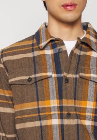 En ung voksen iført en brun, orange, marineblå og hvid ternet skjorte med knapper over en hvid undertrøje, vist fra nakken til brystet.