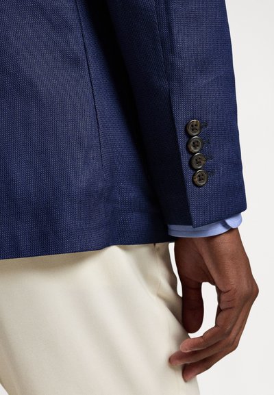 Vyro ranka poilsiui šalia, vilkint mėlynos tamsiai mėlynos spalvos švarką su keturiais mygtukais ant rankogalių, po kuriuo šviesiai mėlyna marškinėliai ir kreminės spalvos kelnės.