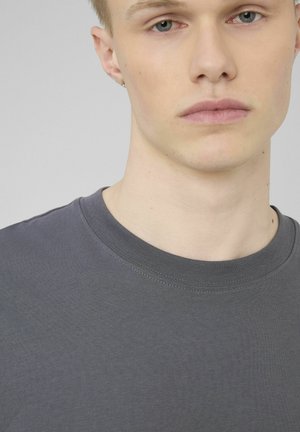 Gros plan d'une jeune personne portant un t-shirt gris uni à col rond sur un fond gris clair.