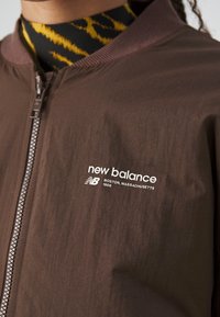 Hnědá zipová bunda s bílým logem "new balance Boston, Massachusetts 1906" na hrudi, nošená přes žlutý a černý vzorovaný top.