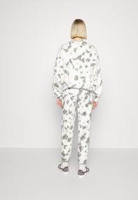 Person med kort blont hår som bär en grå och vit tie-dye sweatshirt och träningsbyxor, står vänd bortåt mot en enkelfärgad vit bakgrund.