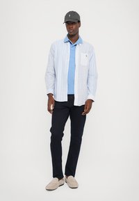 Lichtblauw en witte geruite overhemd met knopen, gelaagd over een blauwe polo, donkere broek en beige instappers. Zwarte pet met logo-accenten.