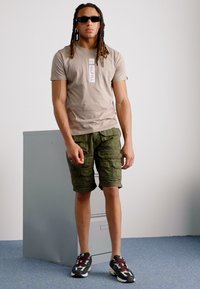 Beige t-shirt med korte ærmer og en hvid label, der står "ALPHA", parret med olivengrønne cargo shorts og mønstrede sneakers.