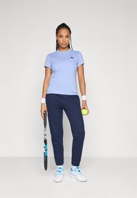 Kvinne i en lys blå t-skjorte, marineblå joggebukser, hvite joggesko og håndleddsbånd holder et tennisrackett og en ball, stående mot en enkel bakgrunn.