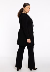 Zwart outfit met een tuniek met een geplooid detail aan de voorkant, gecombineerd met flare broeken. Gladde stof en een getailleerd silhouet. Gestyled met hakken.
