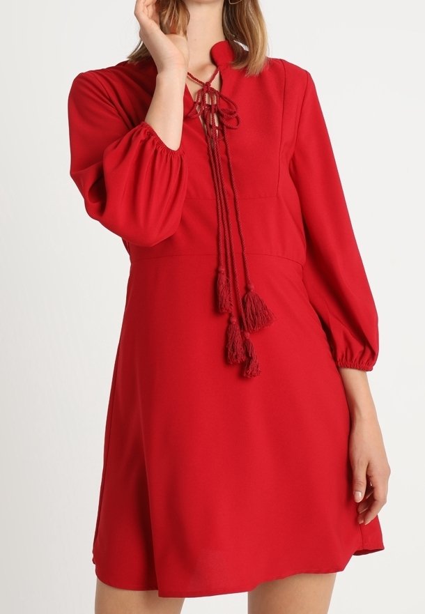 Červené šaty s vysokým výstřihem, dlouhými objemnými rukávy a zavazováním s třásněmi v přední části. Hladký materiál a délka nad kolena.