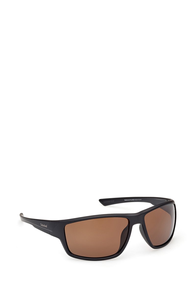 Lunettes de soleil en plastique noir avec des verres teintés marron, présentant un design enveloppant et des branches droites. Détail du logo sur la branche latérale.