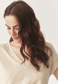 Béžové, texturované tričko s výstřihem do V a krátkým rukávem. Tkanina vypadá měkká a lehká. Model má vlnité tmavě hnědé vlasy, které jsou částečně viditelné.
