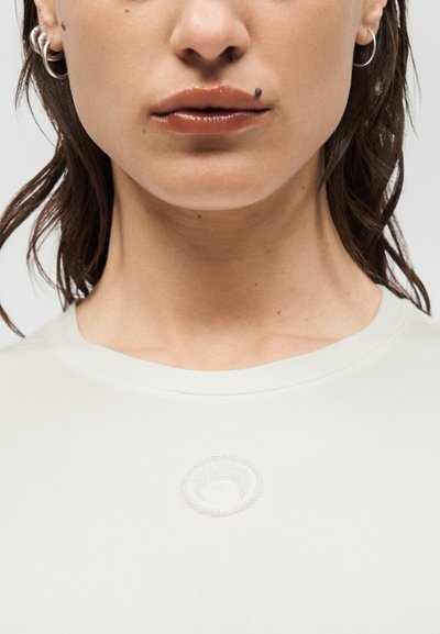 Κοντινό πλάνο ενός ατόμου που φορά λευκό πουκάμισο, με κρίκους στα αυτιά, γυαλιστερά χείλη και μια μικρή μαύρη φακίδα πάνω από το άνω χείλος.