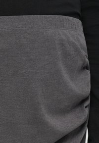 Zone de la taille d'une personne portant un haut noir rentré dans un pantalon gris taille haute avec une ceinture élastique et une poche latérale.