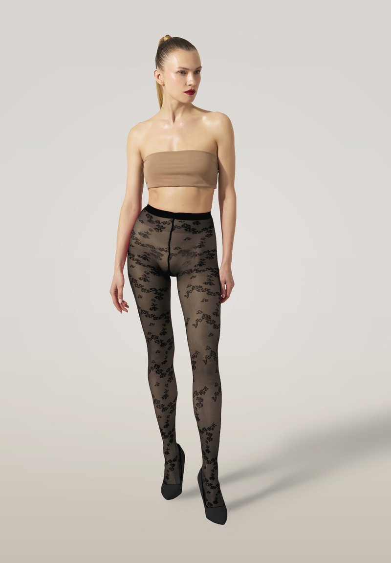 Collant neri con motivi floreali, texture semi-trasparente, vita alta, abbinati a tacchi neri a punta e a un top nudo.