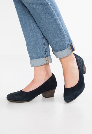 Marineblaue Wildleder-Pumps mit niedrigem Absatz, abgerundetem Zehenbereich, strukturierten Akzenten und dezenten Perforationen, kombiniert mit hochgekrempelten hellen Jeans.