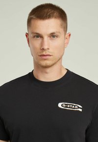 T-shirt en coton noir avec un col rond et un petit logo brodé sur la poitrine gauche. Texture lisse, coupe décontractée.