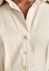 Gros plan d'une chemise beige à col avec trois boutons fermés, montrant la partie supérieure de la poitrine et du cou d'une personne.