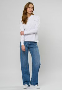 Sieviete ar gariem viļņainiem matiem, valkājot baltu garām piedurknēm polo kreklu, zilas plašu kāju džinsas un baltas sporta kurpes, stāv pret vienkāršu fonu.