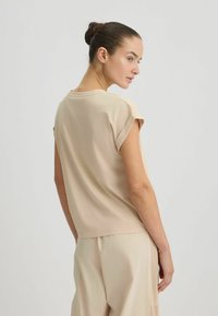 Top beige a maniche corte con una vestibilità rilassata, texture morbida e colletto rotondo. Presenta polsini arrotolati e un orlo leggermente curvato.