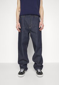 Dark blue denim trousers with a loose fit, featuring a yellow stitching detail and worn with black sneakers.