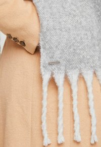 Hall hall koos värviline sall, millel on tekstureeritud kangas ja keerutatud valged äärised otstes, mille külge on kinnitatud helebeez mantel nuppude detailidega.