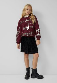 Bordeauxroter Sweatshirt mit metallischem Aufdruck, kombiniert mit einem schwarzen Samtrock mit weißen Pünktchen. Schwarze Schnürstiefel im Combat-Stil.