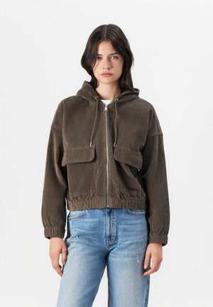 Bruine corduroy jas met capuchon, voorzien van een ritssluiting, twee voorzakken en elastische manchetten. Gemonteerd over lichtblauwe distressed jeans.