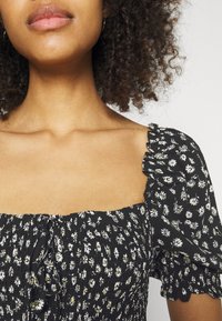 Blouse noire à motifs floraux avec corsage smocké et manches courtes bouffantes. Présente des motifs de marguerites blanches et jaunes, avec des bords de manches volantés.