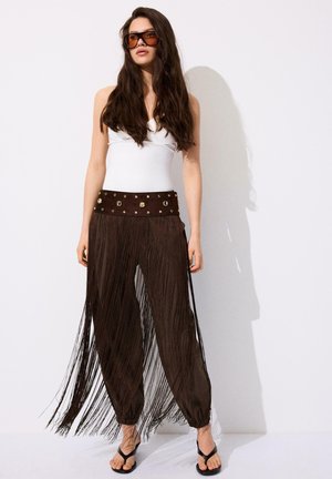 Femme aux longs cheveux foncés portant de grandes lunettes de soleil marron, un haut blanc sans manches, un pantalon marron à franges avec une ceinture cloutée et des sandales noires.