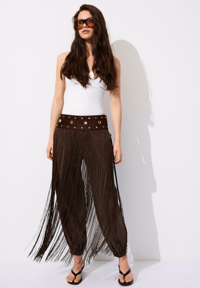 Femme aux longs cheveux foncés portant de grandes lunettes de soleil marron, un haut blanc sans manches, un pantalon marron à franges avec une ceinture cloutée et des sandales noires.