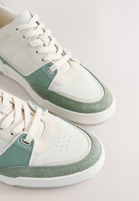 Baskets blanches et vert clair avec un design à lacets, semelle texturée et détails perforés sur la zone des orteils, vues en plongée sous un angle.