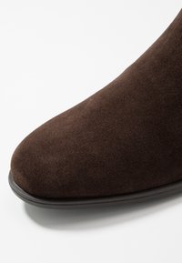 Botín de ante marrón con punta redondeada, que presenta una textura suave y una suela baja de goma negra. El diseño es elegante y minimalista.