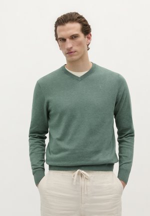 Pulover verde cu decolteu în V, confecționat dintr-un material tricotat moale, cu mâneci lungi și manșete cu model côté, asociat cu pantaloni cu sfoară de culoare deschisă.