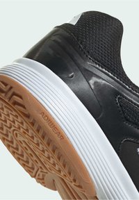 Chaussure de sport noire et blanche avec une semelle extérieure en caoutchouc marron texturée affichant le logo Adidas Adiwear et un petit swoosh Nike sur le panneau noir du talon.