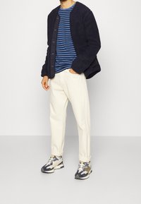 Námořnická fleecová bunda s knoflíky, proužkovaná modro-bílá košile, krémové kalhoty a vícebarevné tenisky s maskáčovým vzorem.