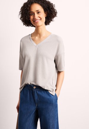 Kvinde med krøllet mørkt hår, smilende, iført en lysegrå V-hals kortærmet sweater og løse blå cowboybukser, står med den ene hånd i lommen.