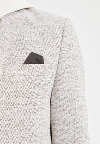Blazer grigio chiaro con tessuto testurizzato, completo di un fazzoletto da taschino nero con pois rossi e sottili dettagli di cucitura.